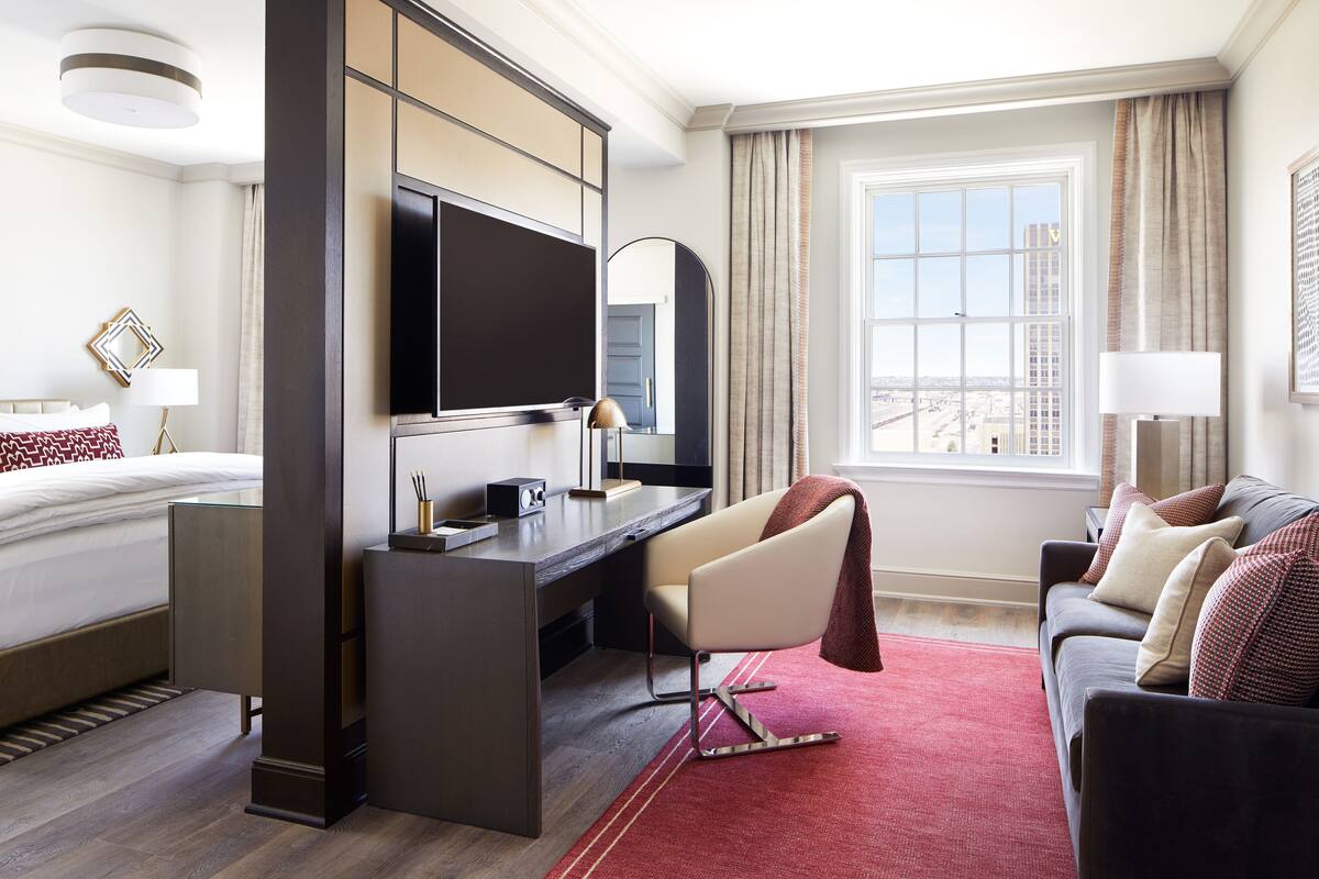 A modern hotel room with a bed, desk, and sofa, a large window with a city view, and a flat-screen TV.
