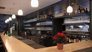 Un espace bar doté de plusieurs étagères remplies de bouteilles d’alcool et de verres, d’un long comptoir et de luminaires suspendus.