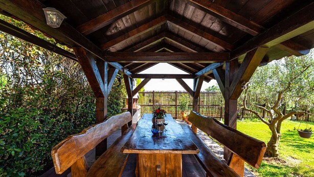 Un pavillon en bois avec une table et des bancs, entouré de verdure et délimité par une clôture.