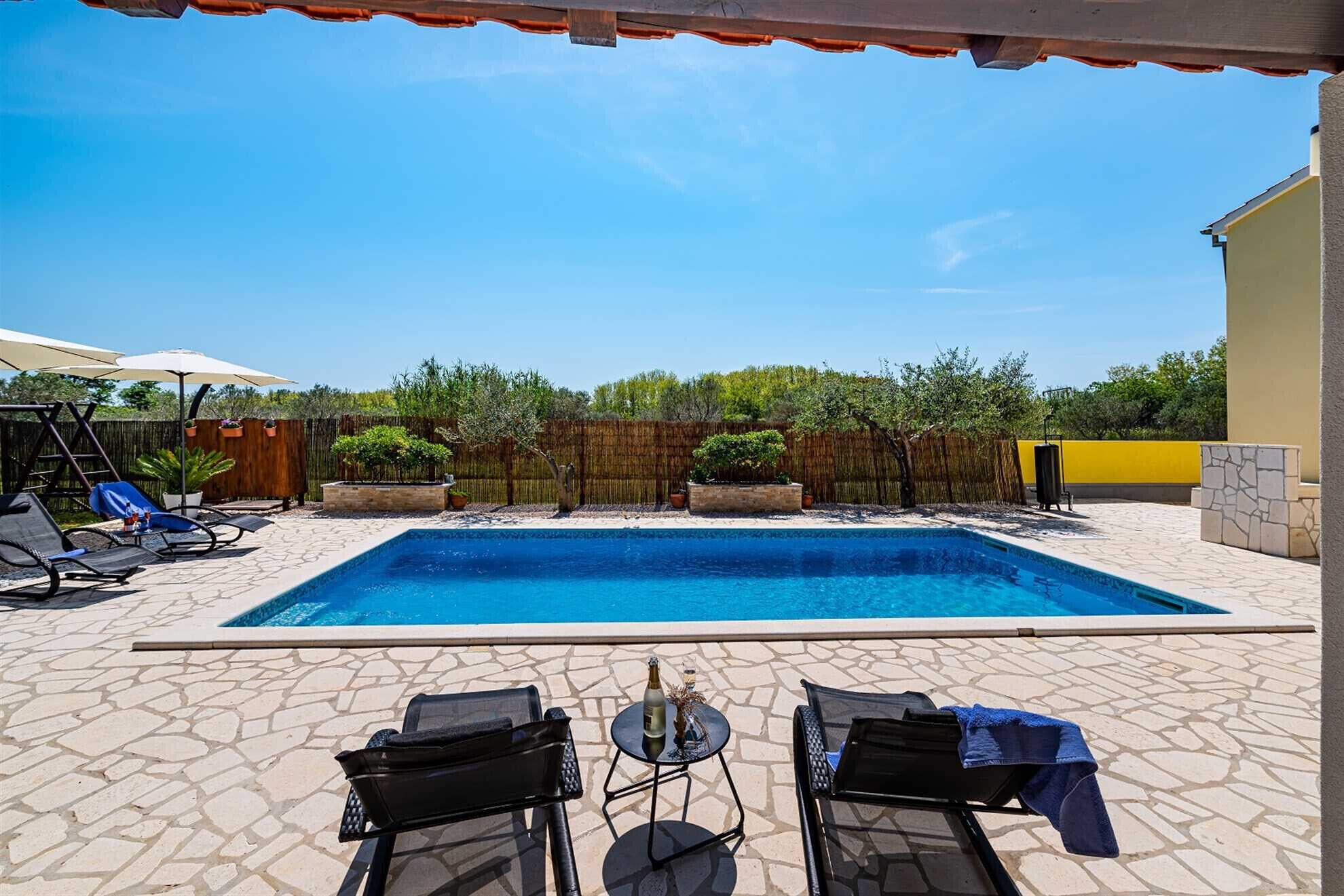Piscina retangular com um deck ladrilhado, rodeada por espreguiçadeiras e uma mesa pequena com uma garrafa e um copo.