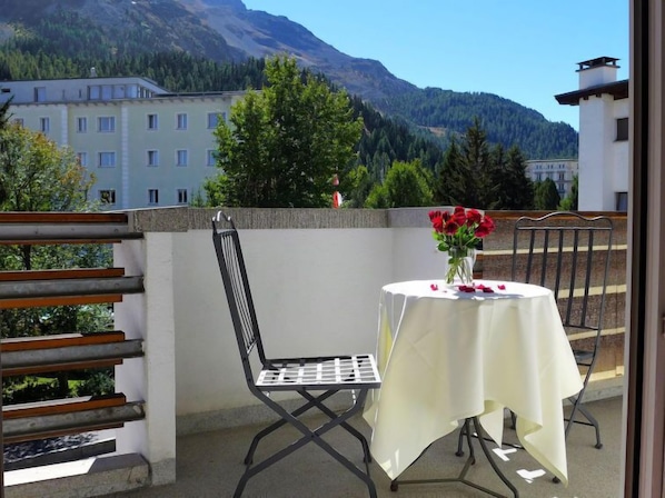 A balcony with a table and chairs, a vase with red flowers, and a mountain view.