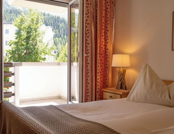 A hotel room with a bed, a nightstand, a lamp, and a view of a forest through a sliding door.