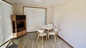 Ein kleiner, gut beleuchteter Raum mit einer Holz-Unterhaltungseinrichtung, einem runden Tisch mit vier Stühlen und einem großen Fenster mit Jalousien.