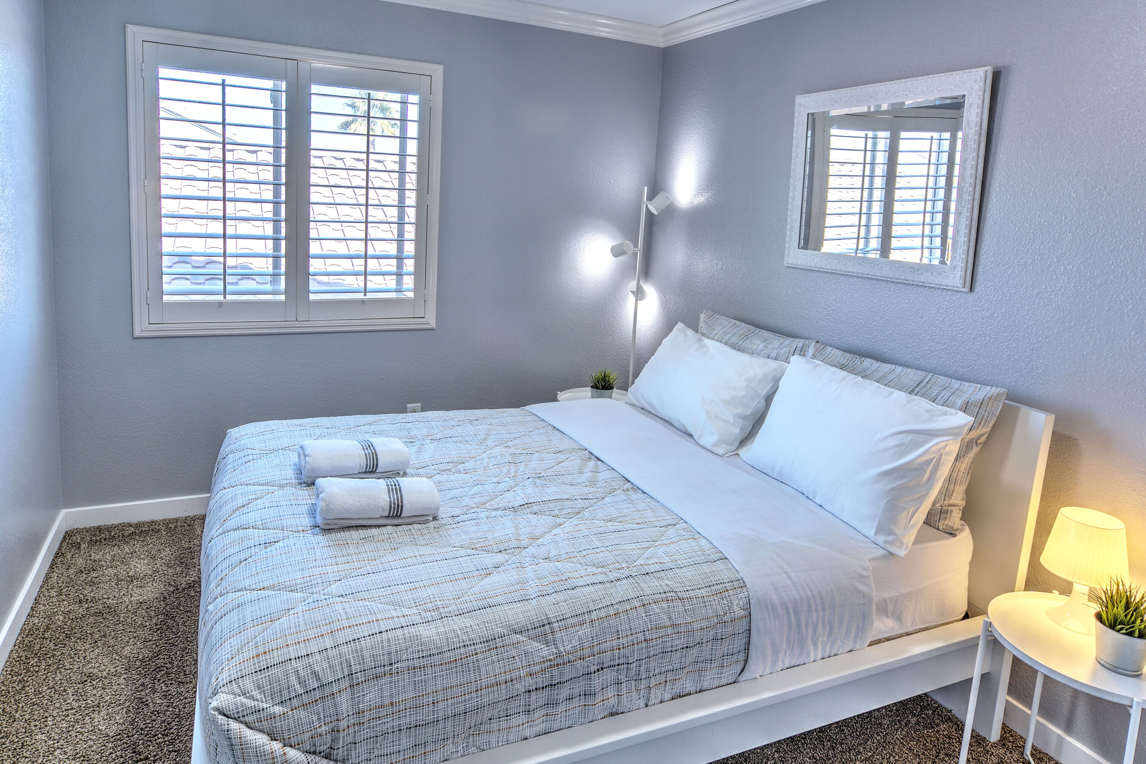A neatly made bed with white linens and a striped blanket, a bedside table with a lamp and a small plant, and a window with blinds.