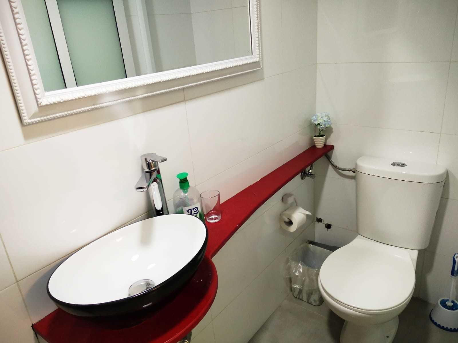 Un baño con lavamanos blanco, encimera roja, inodoro y espejo sobre el lavamanos.