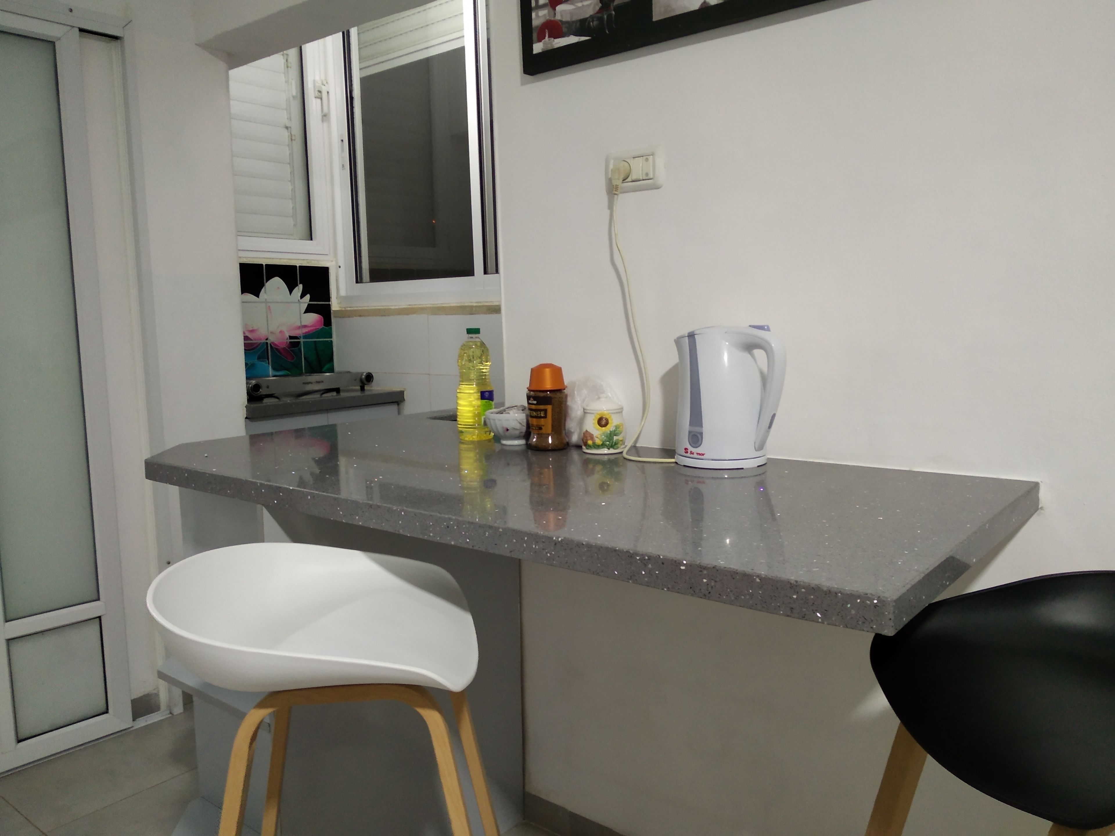 A kitchen with a countertop, a white sink, a black chair, a white chair, a kettle, a bottle of oil, and a small container.
