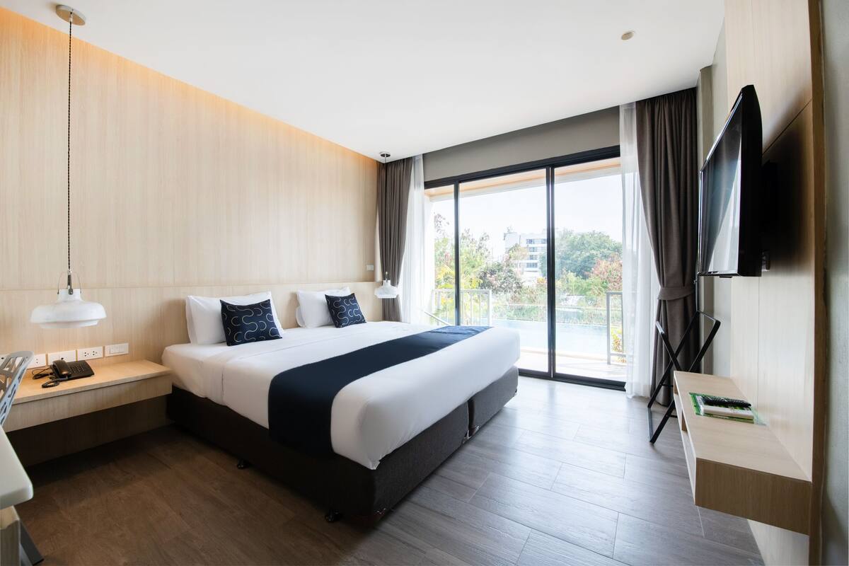 Ein modernes Hotelzimmer mit einem großen Bett, einem Schreibtisch mit Telefon, einem an der Wand montierten Fernseher und einem Balkon mit Blick auf die Stadt.