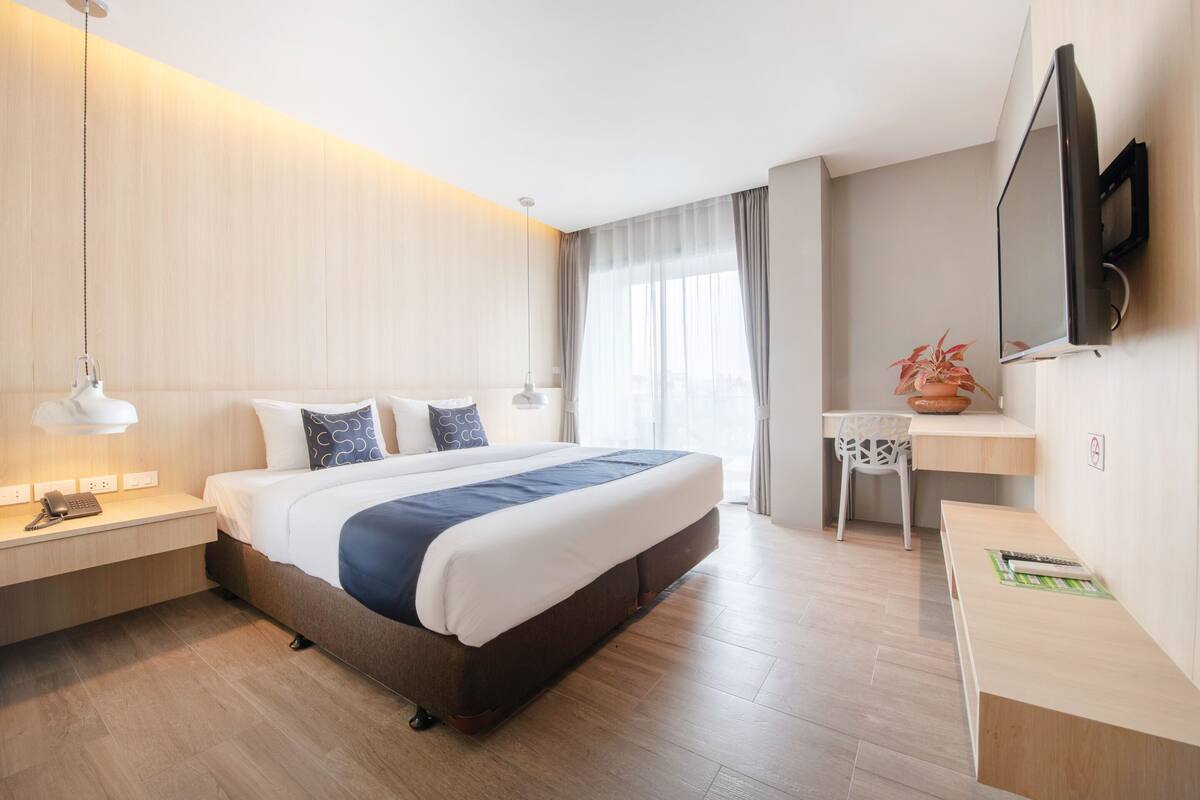 Ein modernes Hotelzimmer mit einem großen Bett, Nachttischen, einem Schreibtisch, einem Sessel sowie einem Fernseher und einem Fenster mit Vorhängen.