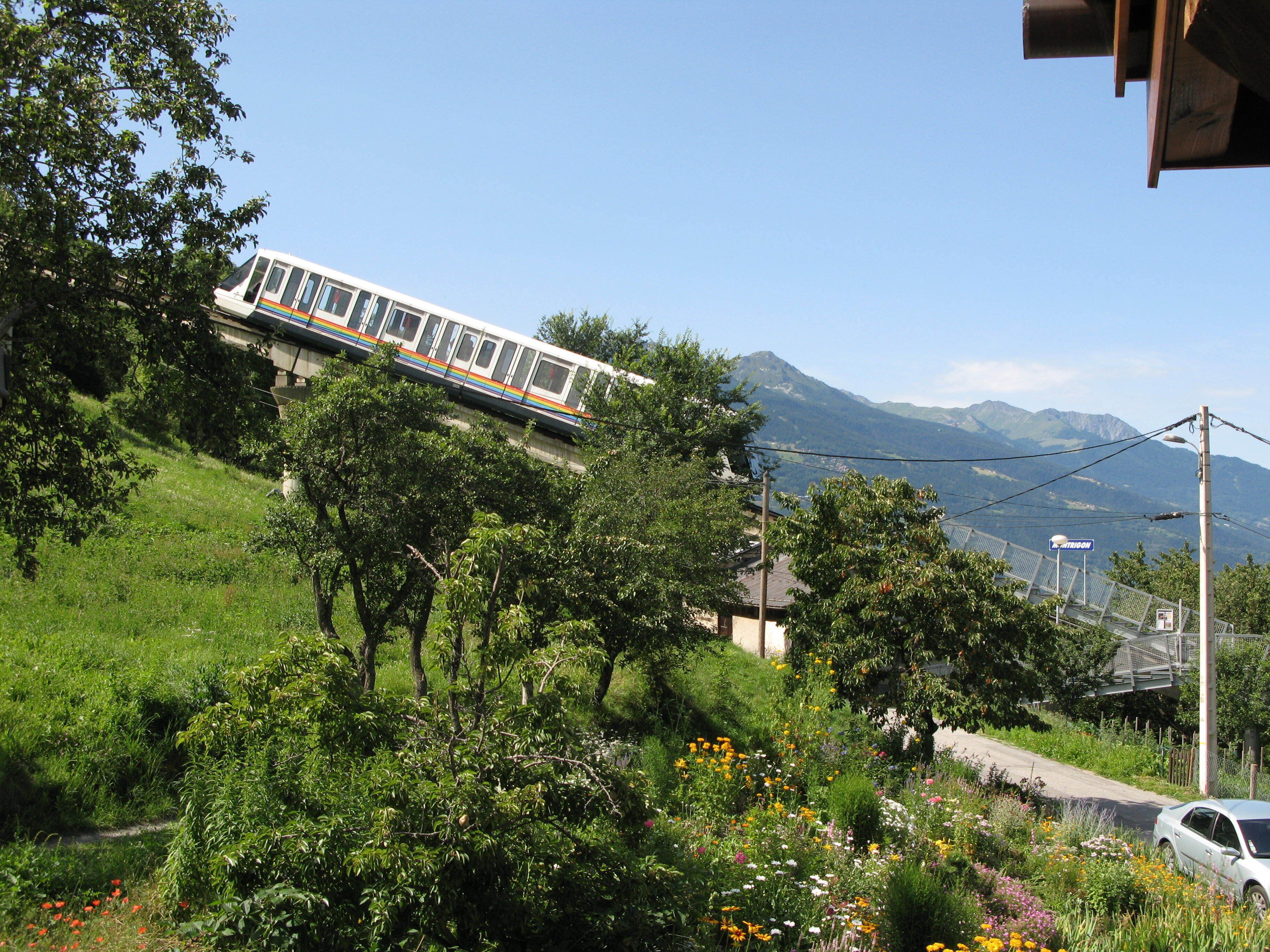 Un train circule sur une voie ferrée, avec en arrière-plan un paysage montagneux, le tout étant entouré de verdure, et une voiture est garée à proximité.