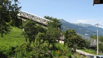 Un train circule sur une voie ferrée, avec en arrière-plan un paysage montagneux, le tout étant entouré de verdure, et une voiture est garée à proximité.
