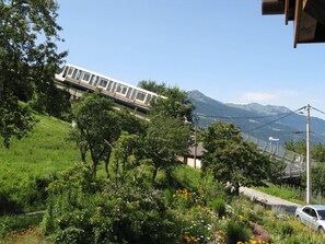 Un train circule sur une voie ferrée, avec en arrière-plan un paysage montagneux, le tout étant entouré de verdure, et une voiture est garée à proximité.