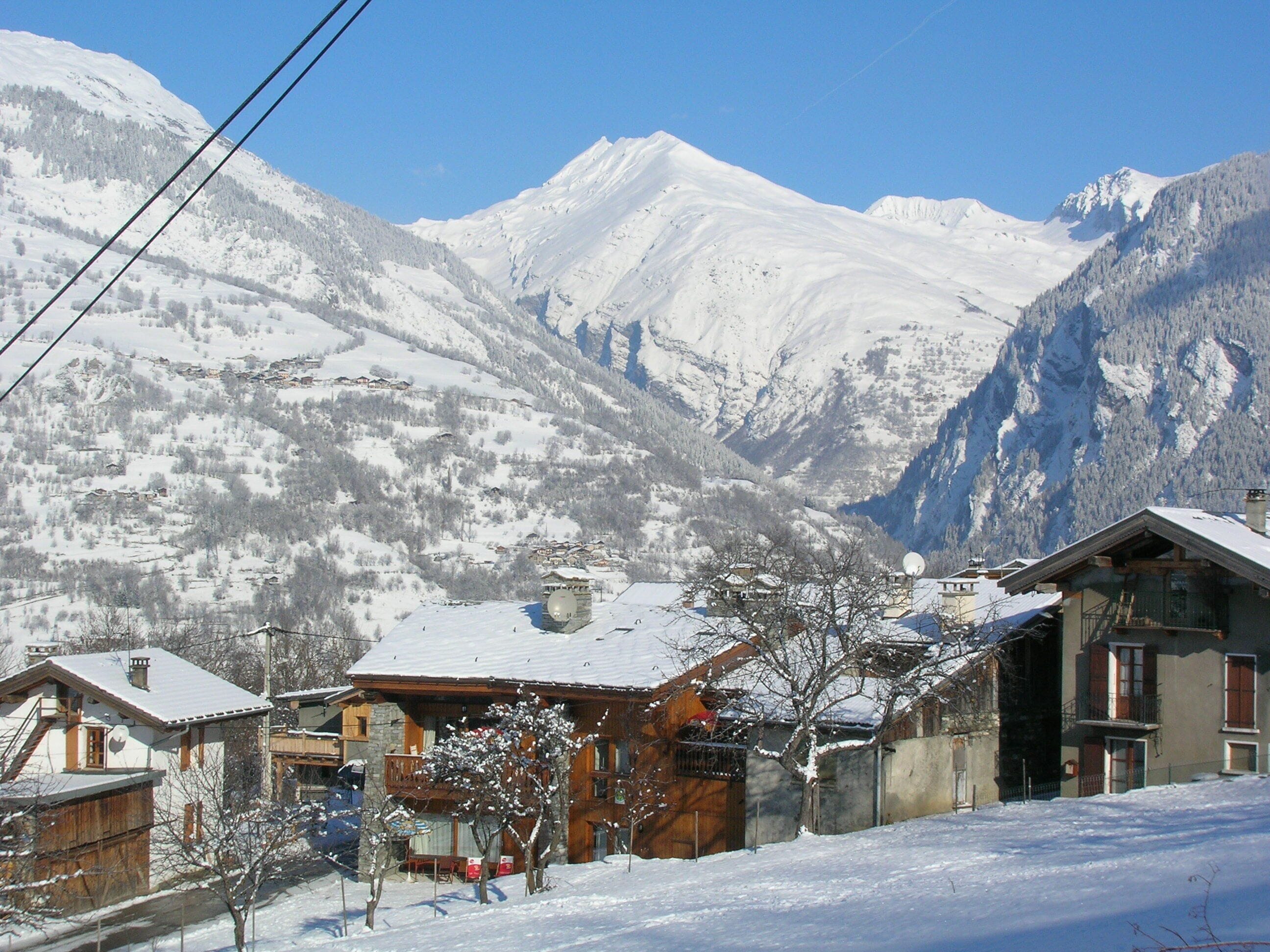 A snow-covered mountain village with traditional wooden houses, a clear sky, and a view of the surrounding peaks.