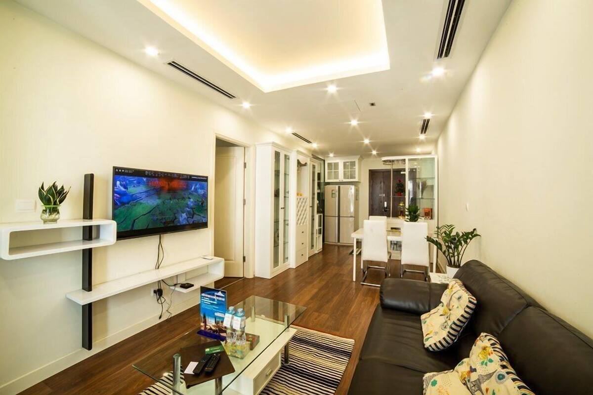 A modern living room with a flat-screen TV, a glass coffee table, a sofa, and a kitchen area in the background.