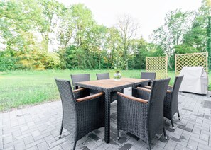 An outdoor patio with a wooden table and wicker chairs, surrounded by greenery and a wooden fence.