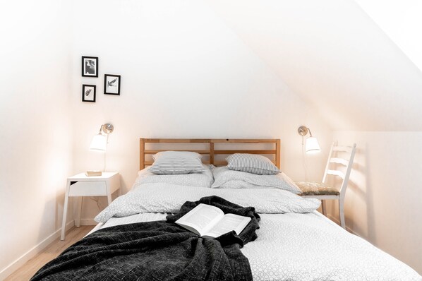 A bedroom with a sloped ceiling, a bed with a black blanket and an open book, two bedside tables with lamps, and a chair.