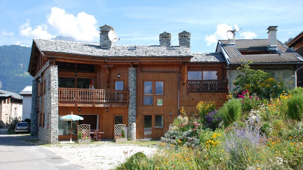 Un edificio in legno a due piani con un balcone, pareti in pietra e un giardino con diverse fioriture.