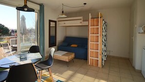 Una habitación compacta con una cama litera, una mesa de comedor con sillas, una pequeña cocina y un ventanal con vistas al exterior.