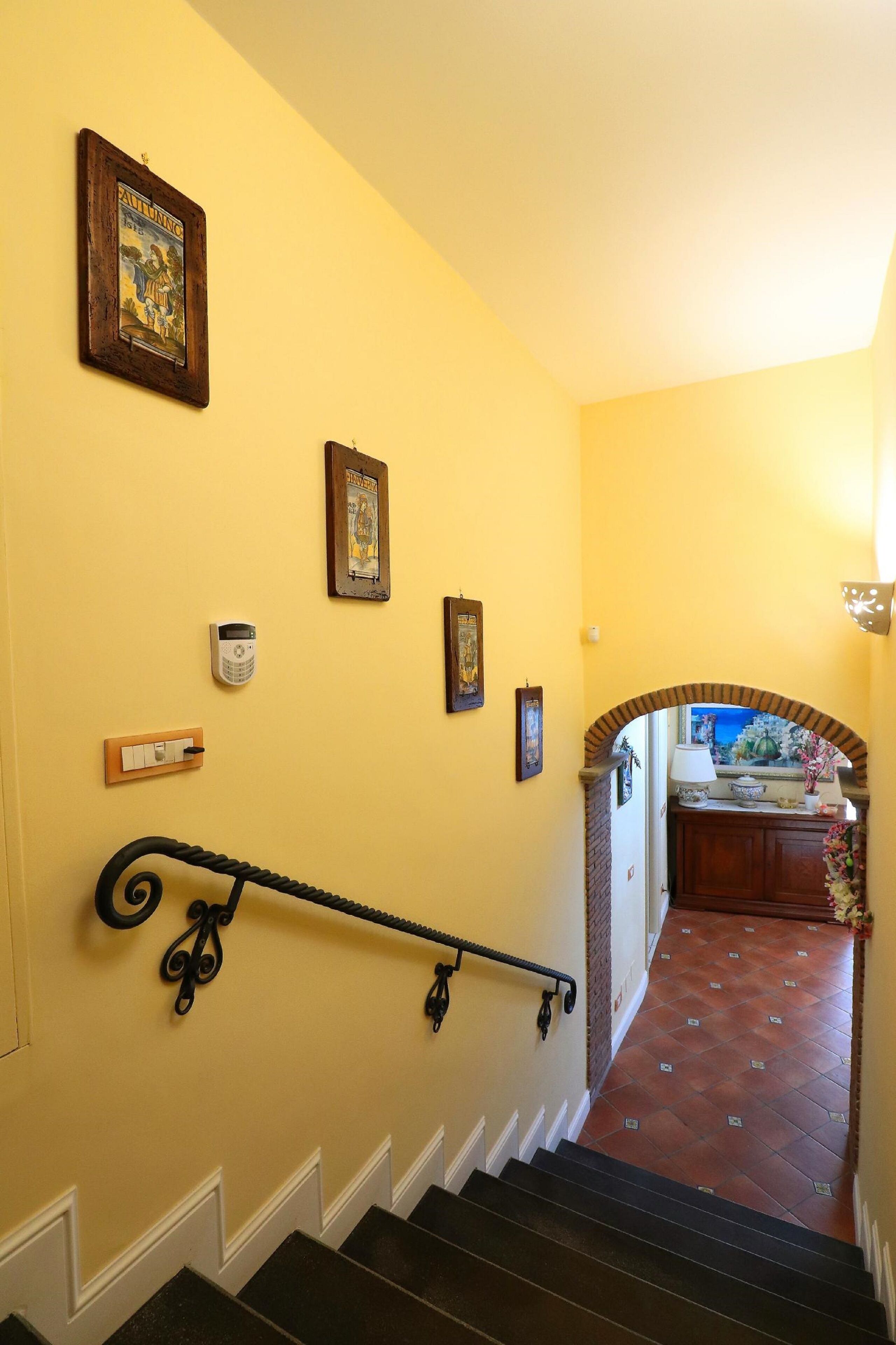 A staircase with a handrail, a wall-mounted light switch, and a door leading to a room with a tiled floor and a decorative mirror.
