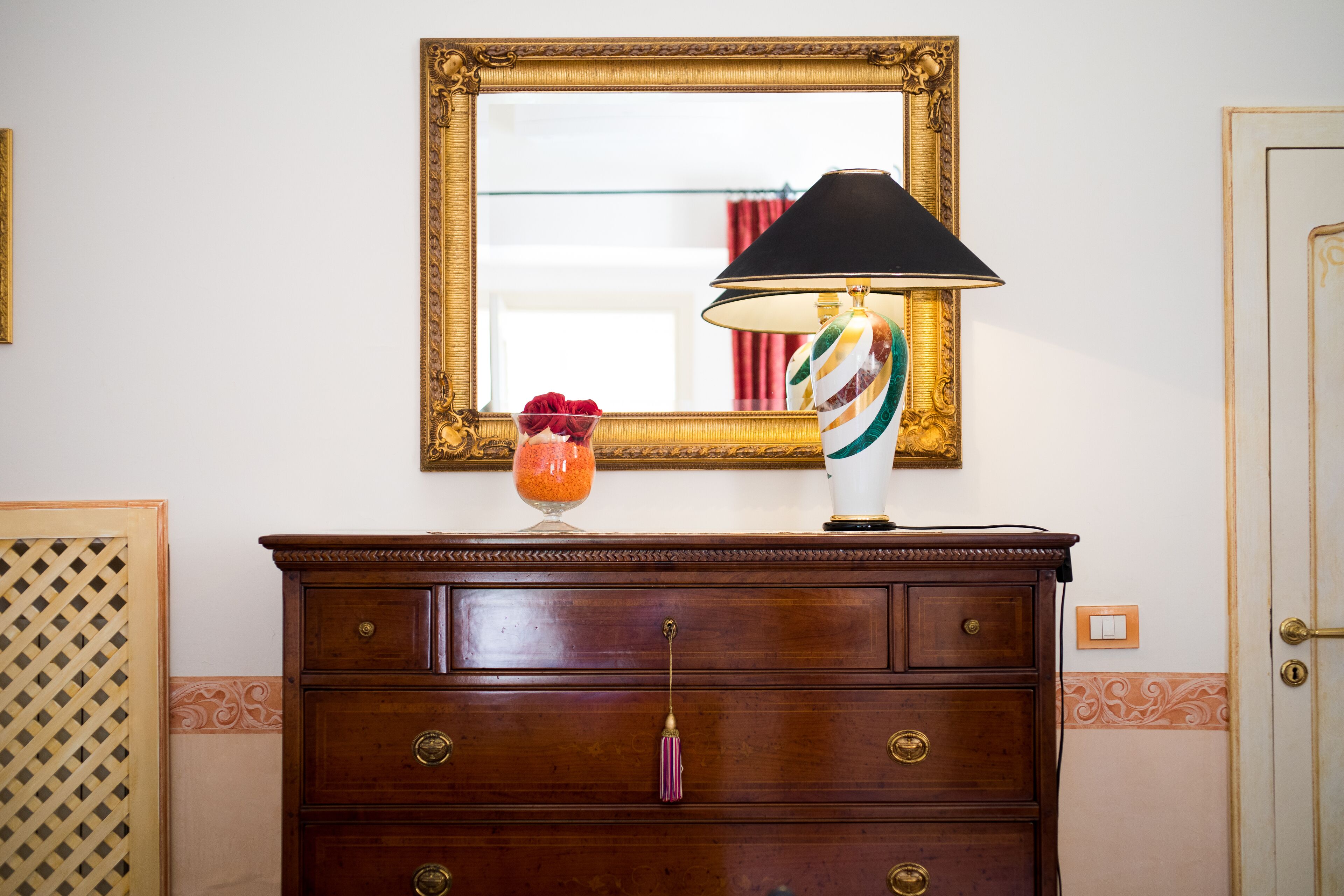 A wooden dresser with a mirror above it, a lamp with a striped lampshade, and a vase with red flowers.