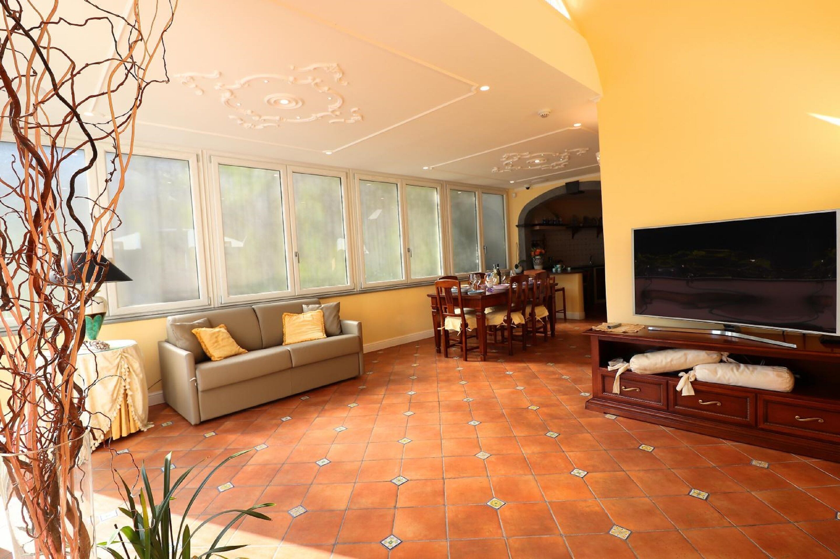 A living room with a sofa, dining table, chairs, a TV, and large windows.