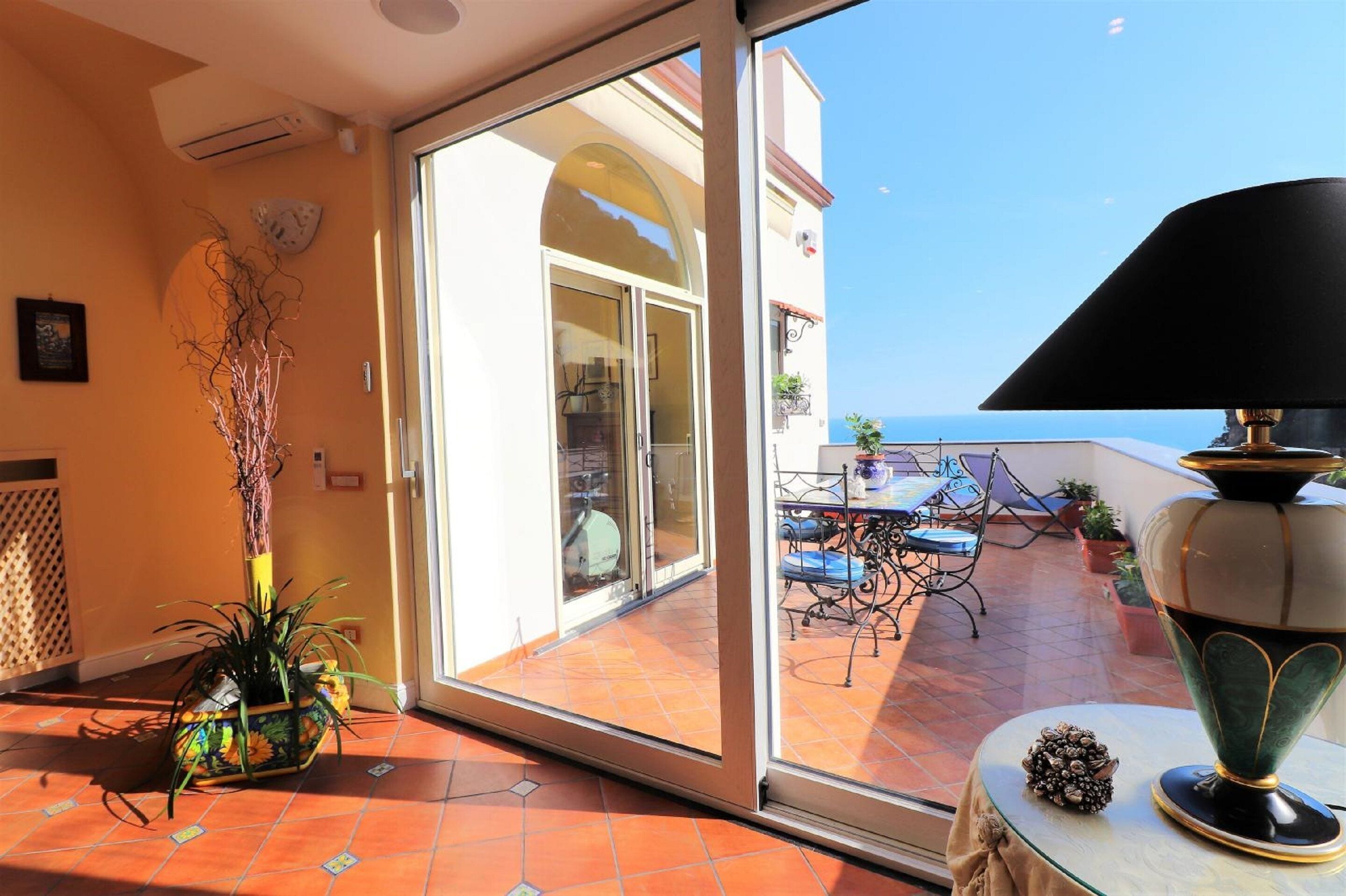 A balcony with a table and chairs, a potted plant, and a view of the sea.