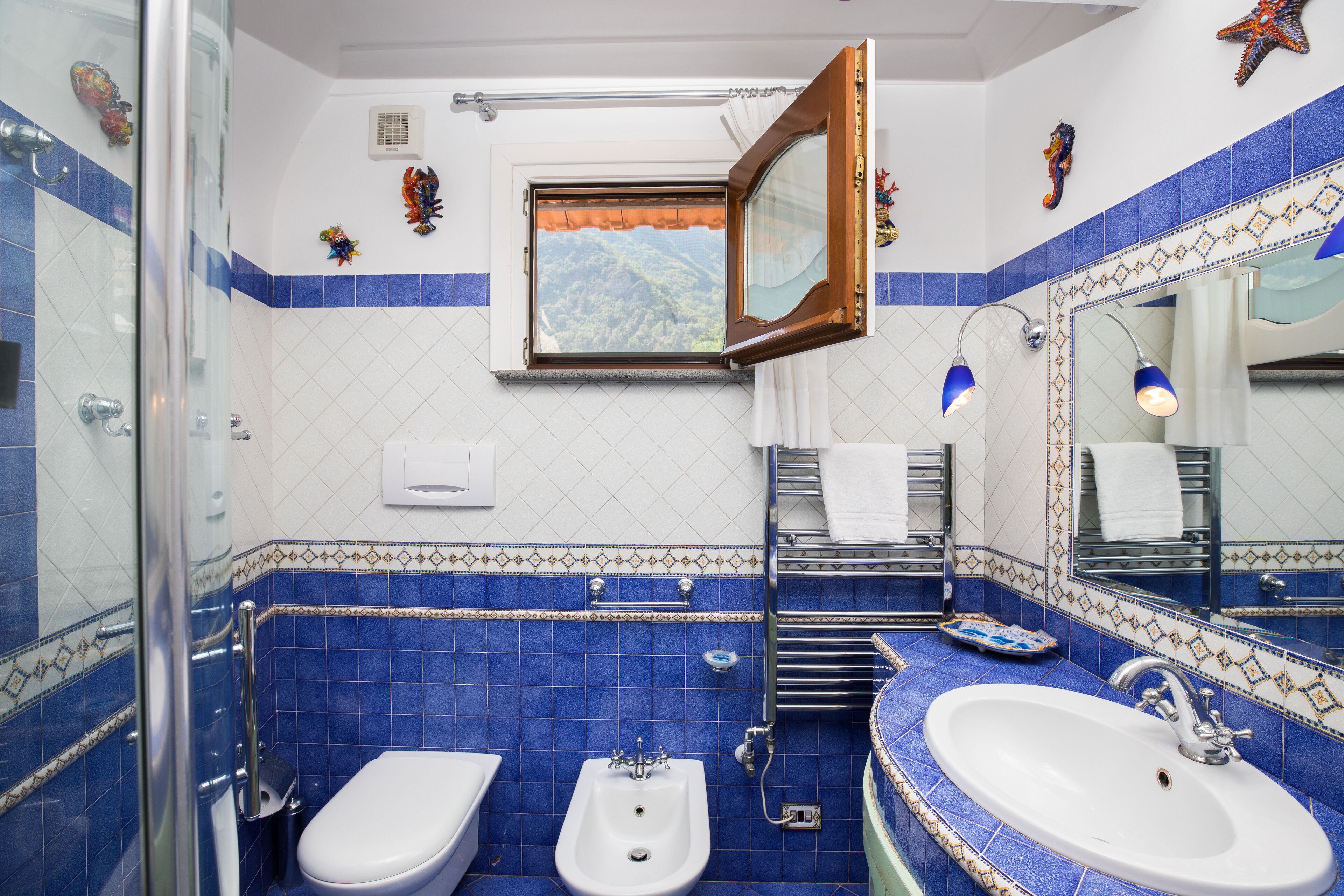 A bathroom with two sinks, a bidet, a shower, and a window with a view of a mountainous landscape.