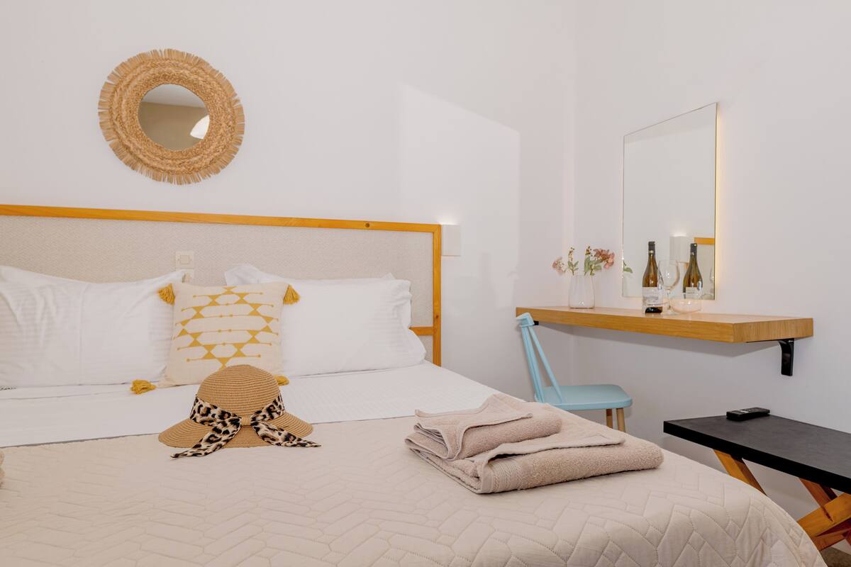 A neatly arranged hotel room with a bed, a chair, a desk, and a shelf with toiletries.