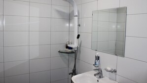 Baño con azulejos blancos, ducha, lavamanos, espejo y artículos de aseo.