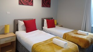 Habitación de hotel con dos camas, cada una con un cojín rojo y una lámpara amarilla. Hay dos cuadros de rosas rojas en la pared.