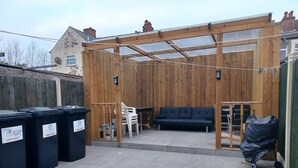 Un espacio de patio cubierto con una pérgola de madera, un sofá negro y una silla blanca. Hay contenedores negros señalizados para la recogida de residuos.