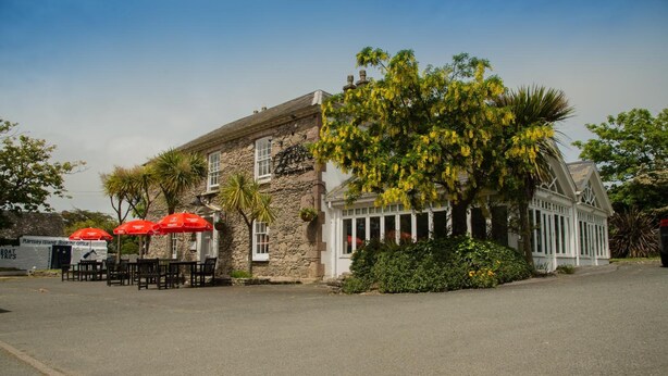 A stone building with a large tree in front, outdoor seating with red umbrellas, and a clear sky. - The Grove Hotel (Haverfordwest)