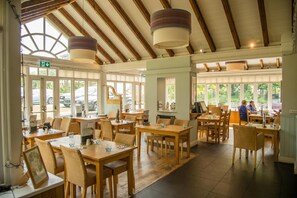 Un vaste espace de restauration intérieur, doté de tables et de chaises en bois, de grandes fenêtres et d’un haut plafond avec des poutres apparentes.