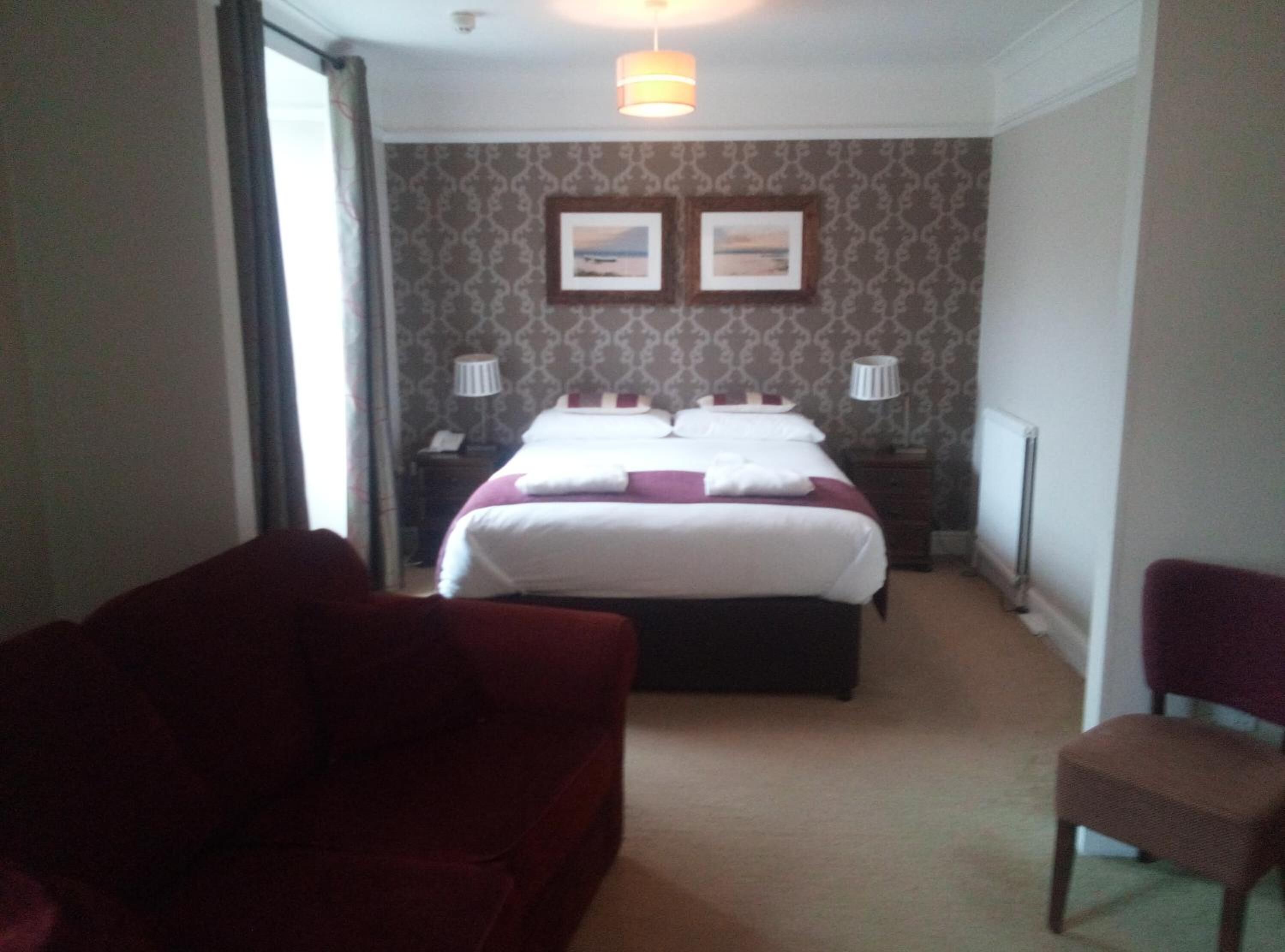 Una habitación de hotel con una cama, dos mesitas de noche, una lámpara, una silla y un sofá rojo.