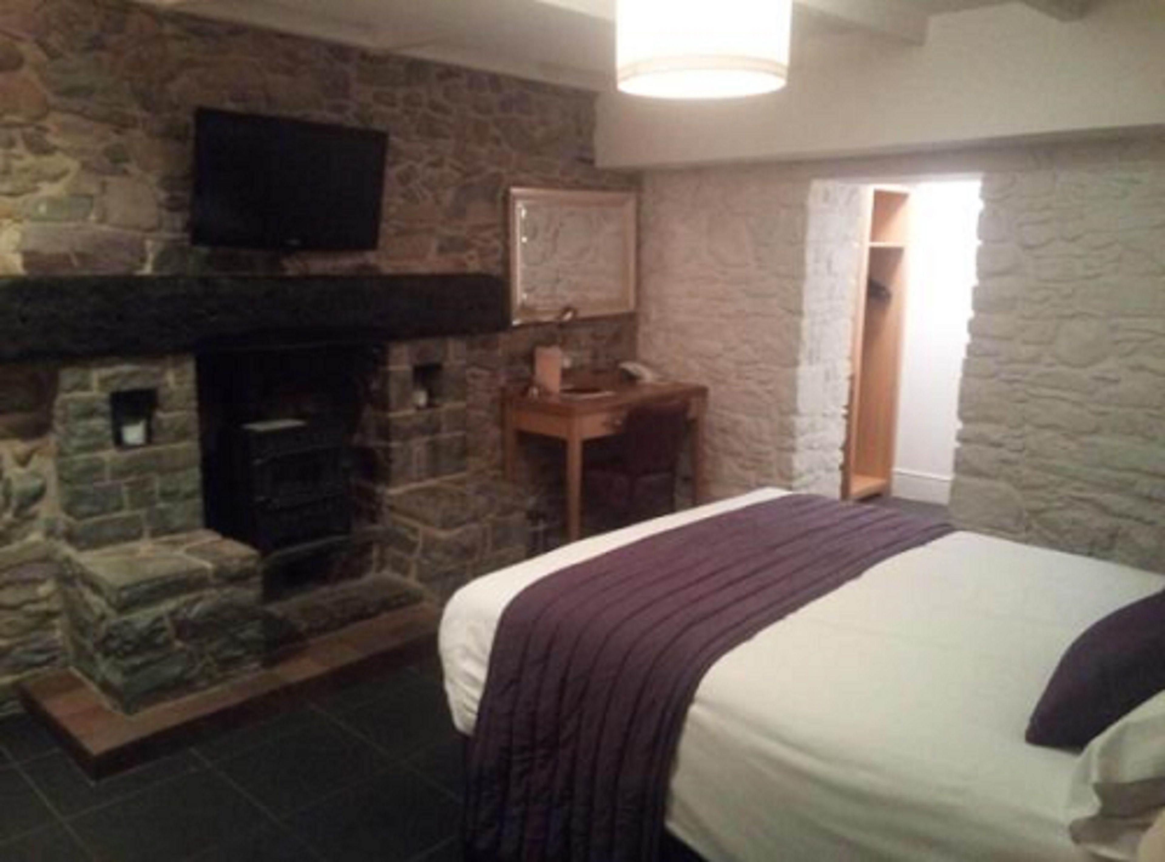 A bedroom with a stone fireplace, a bed with a purple blanket, and a desk with a chair.