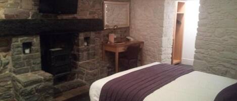 A bedroom with a stone fireplace, a bed with a purple blanket, and a desk with a chair.