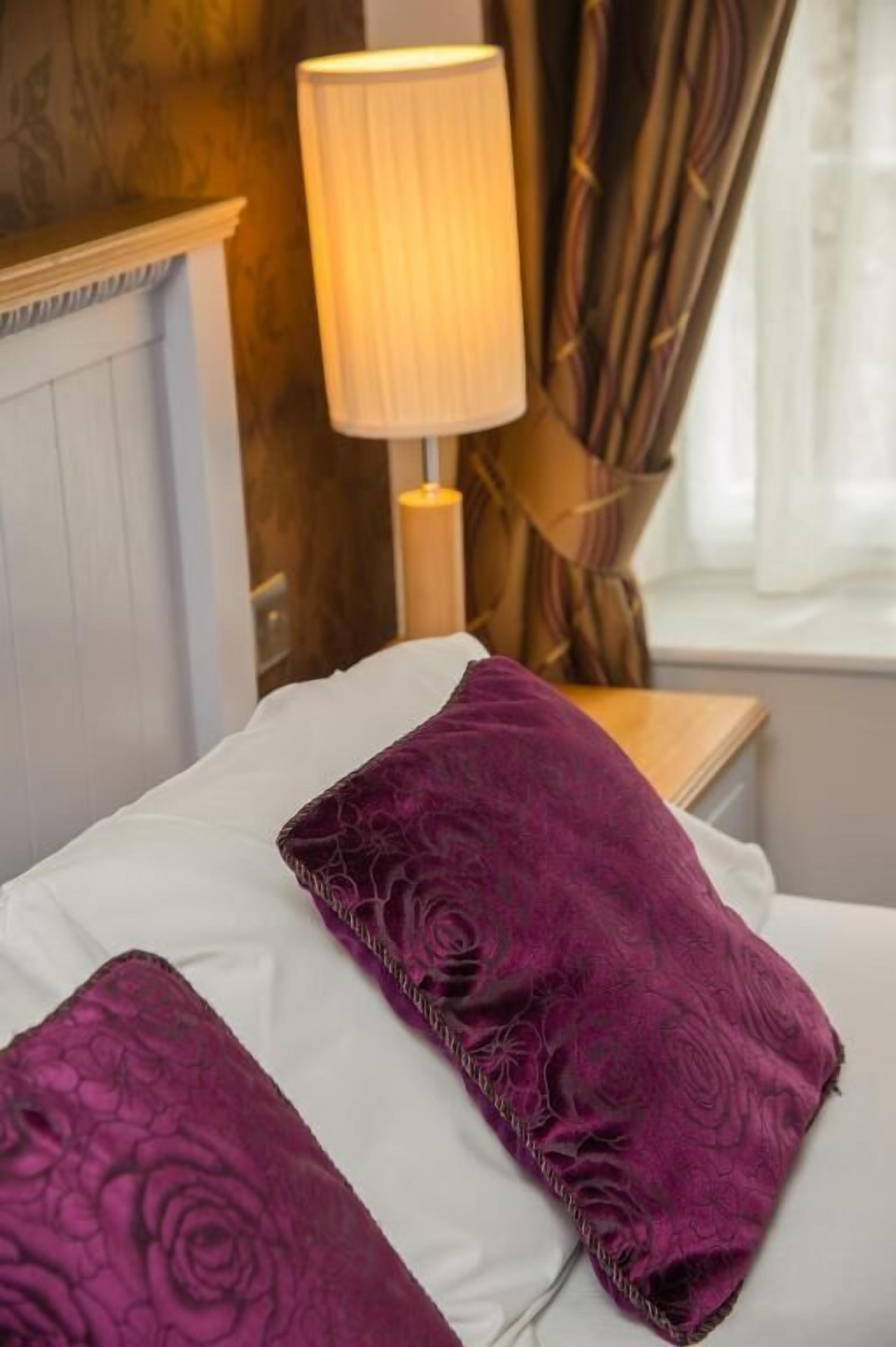 A bed with white and purple pillows, a bedside lamp, and a window with curtains.