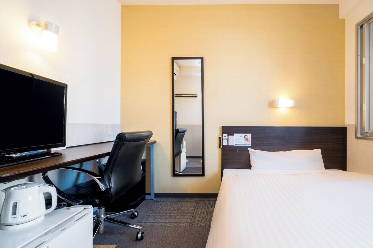 A hotel room with a bed, a desk with a TV, a chair, a coffee maker, a mirror, and a window.