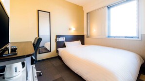 Hotellihuone, jossa on sänky, työpöytä tietokoneella, tuoli, kahvinkeitin ja ikkuna, jossa on sälekaihtimet.