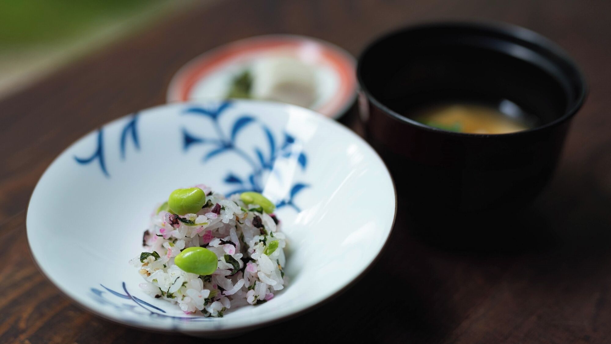 A bowl of rice with green beans and a small cup of soup on a wooden table.