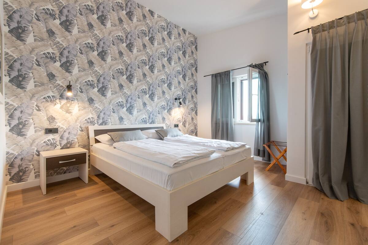 Un dormitorio con cama, mesitas de noche, una ventana con cortinas y una pared con estampado de hojas.
