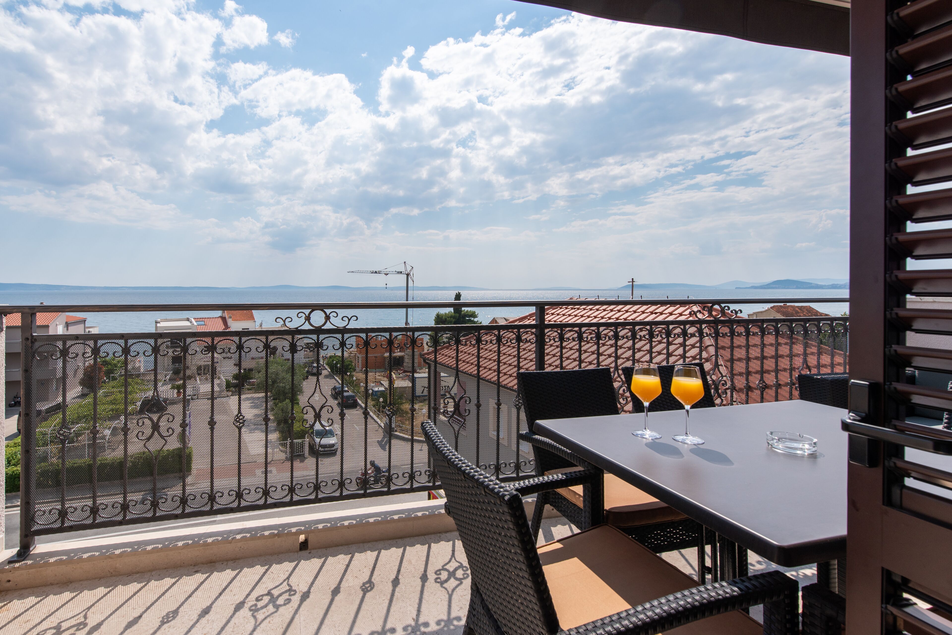 Un balcon avec une table et des chaises, deux verres de jus d’orange et une vue sur la mer et le paysage urbain.
