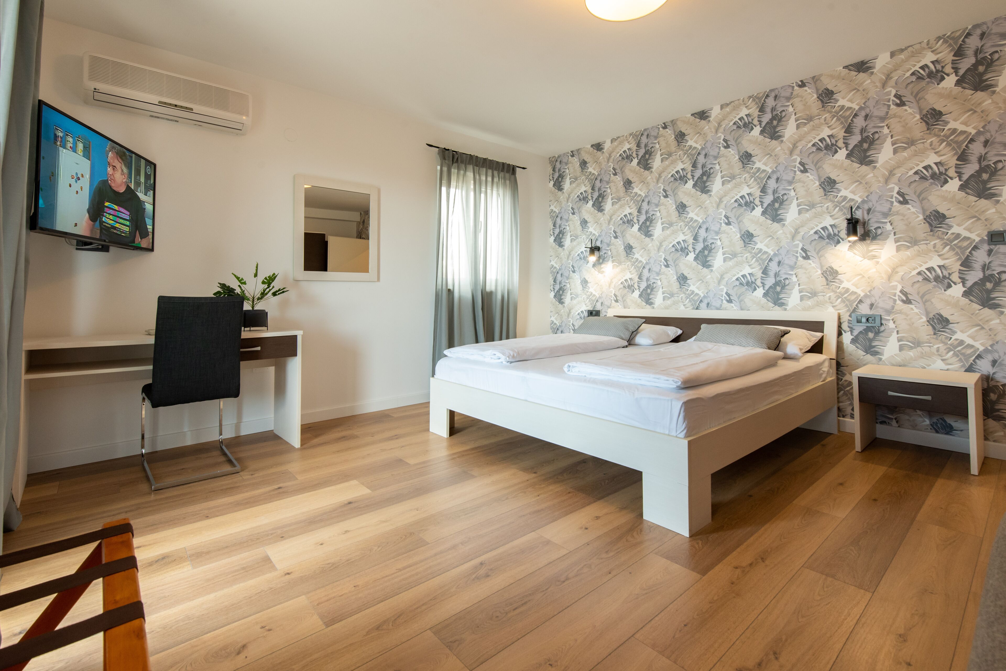 A modern bedroom with a white bed, a desk with a chair, a small bedside table, and a wall-mounted TV.