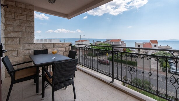 Un balcon avec une table et des chaises, un mur en pierre, une vue sur la mer et une rue où circulent des voitures.