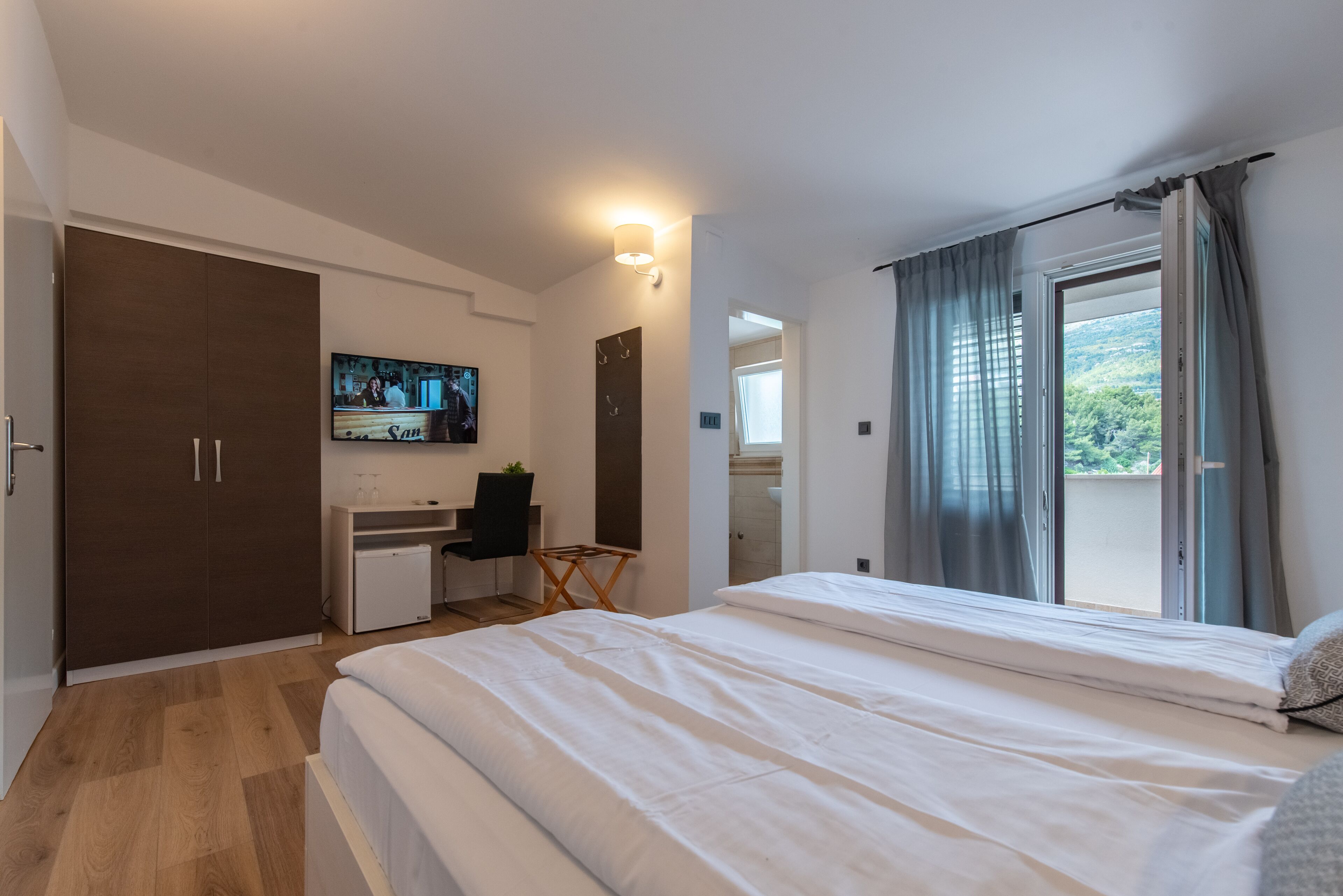 A bedroom with a bed, a desk, a TV, and a balcony with curtains.