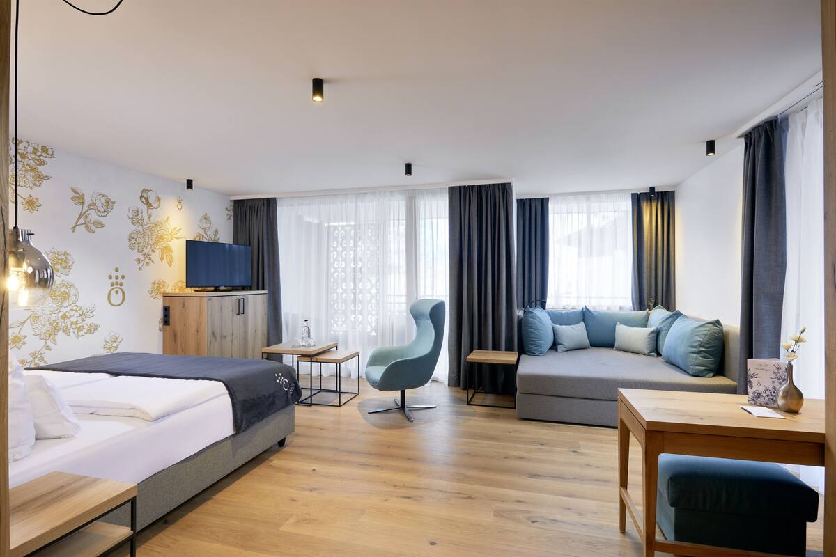 A modern hotel room with a bed, a sofa, a desk, and a chair. There is a TV on a cabinet, and the room has wooden flooring and a window with curtains.