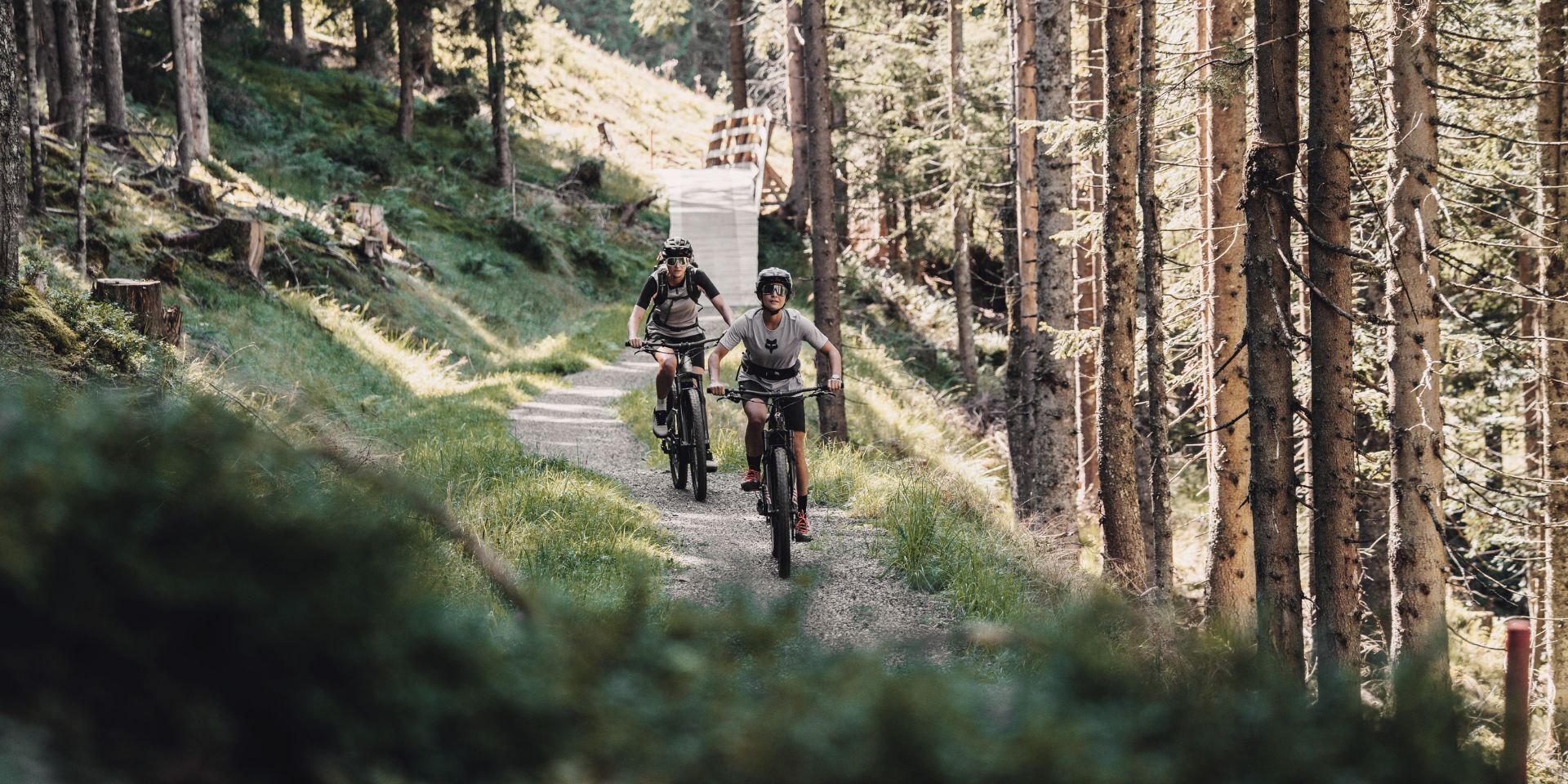 Due ciclisti su un sentiero boschivo, con un ponte sullo sfondo.