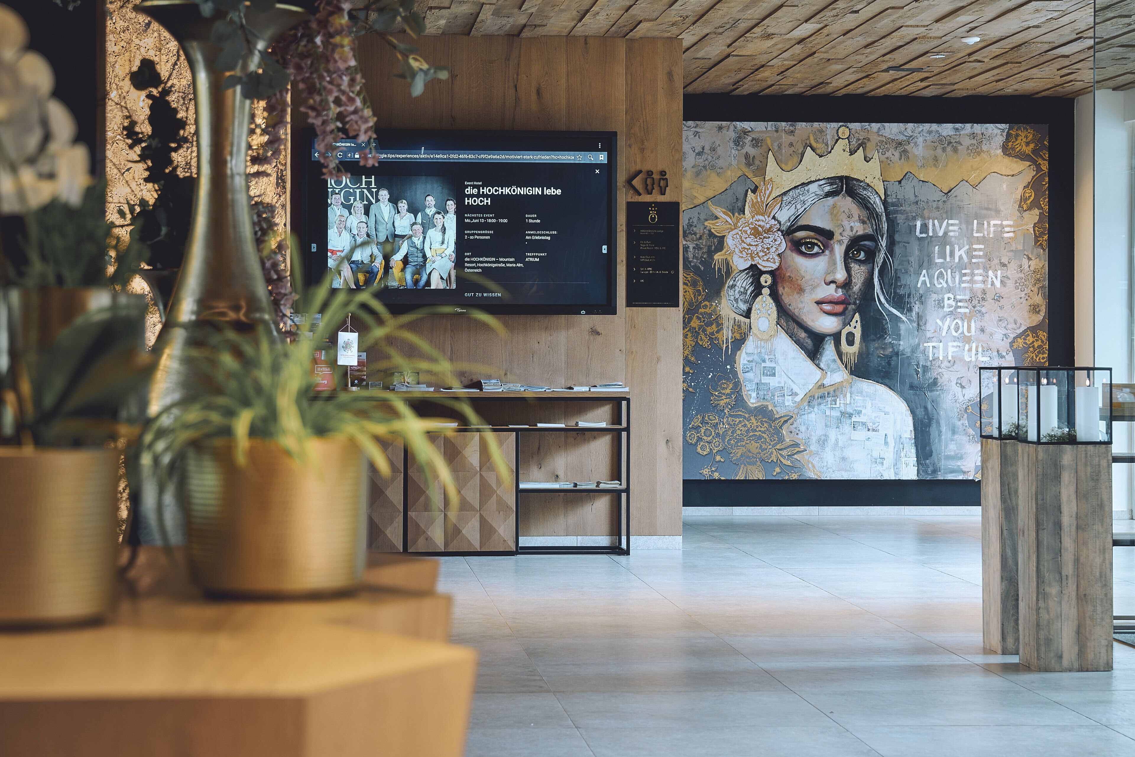 Zona de recepción con un escritorio de madera, plantas en macetas, una pantalla de televisión y un mural con una corona y texto motivacional.