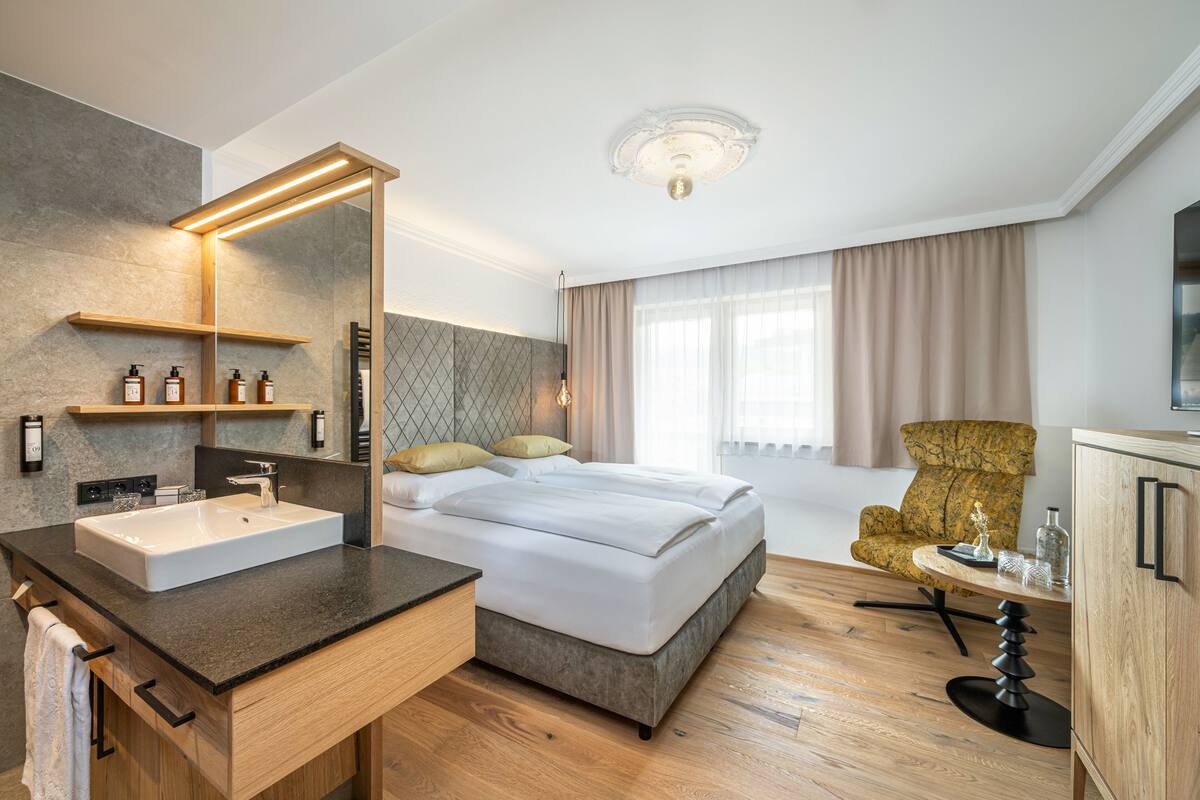 A modern hotel room with a large bed, a built-in vanity with a mirror, a wooden floor, and a large window with curtains.