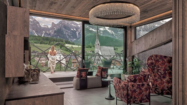 A room with a wooden ceiling, a large chandelier, and a view of mountains and a church. There are two sofas and a table with chairs. - die Hochkönigin - Mountain Resort (Maria Alm am Steinernen Meer)