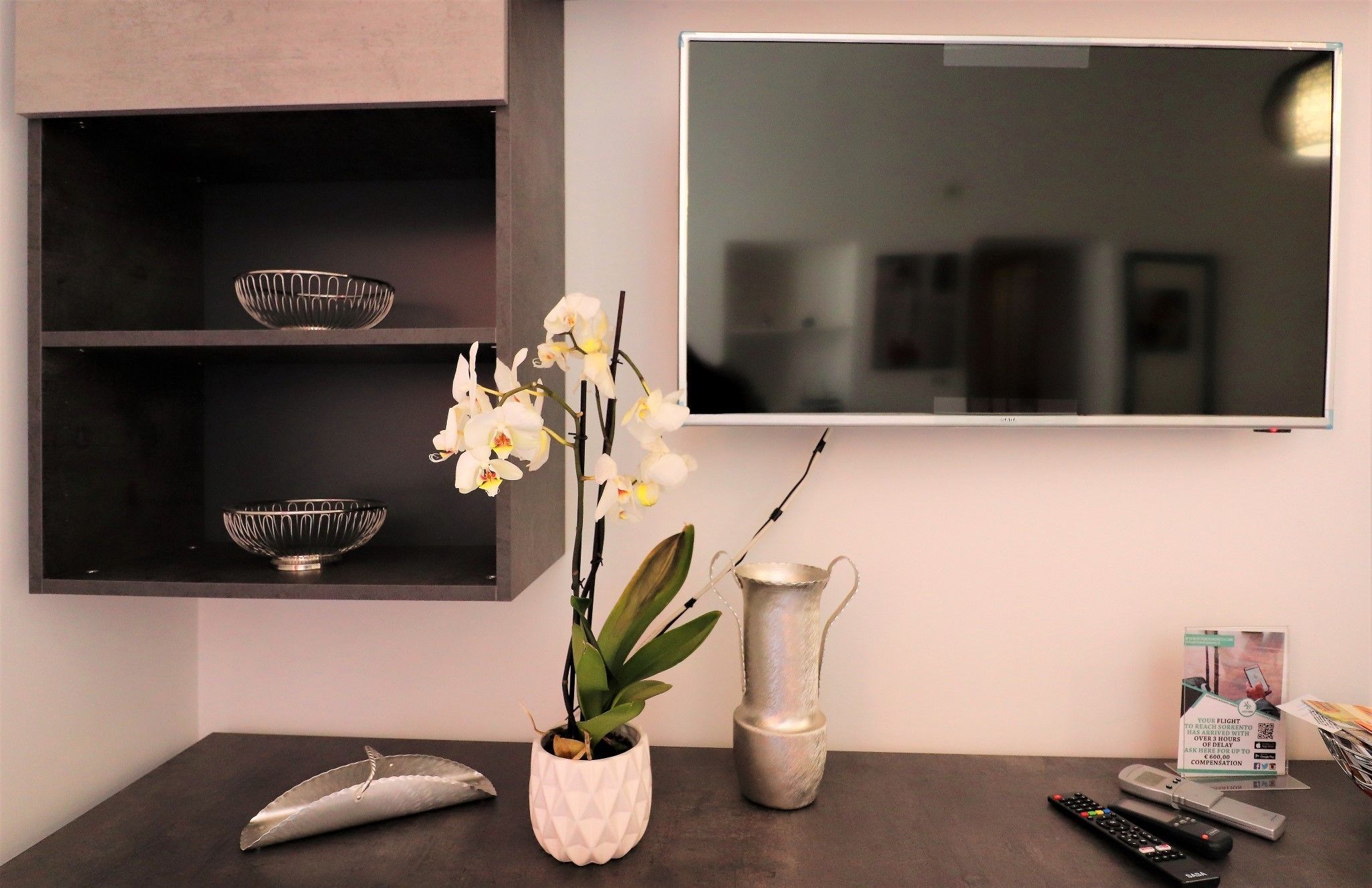 Una habitación moderna con una televisión, una estantería con cuencos decorativos, una planta de orquídea blanca, un florero plateado y un control remoto sobre una superficie oscura.